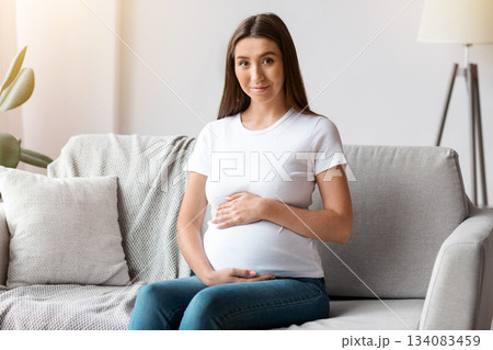 Happy Expectation. Portrait Of Beautiful Pregnant Woman Sitting On Couch At Home, Millennial Expectant Female Embracing Her Belly And Smiling At Camera While Relaxing On Sofa In Living Room 134083459