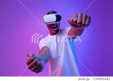 Young african american man playing video games in VR-glasses, excited black millennial guy experiencing virtual reality while standing in neon light over purple gradient background, copy space Young african american man playing video games in VR-glasses, excited black millennial guy experiencing virtual reality while standing in neon light over purple gradient background, copy space 134083482