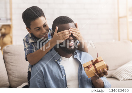 Father's Day. Happy son congratulating his afro dad, covering his eyes from behind and giving present at home, free space 134083490