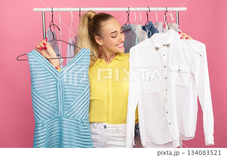 Portrait of young smiling lady standing in clothes shop choosing stylish outfit, holding hangers with dress and shirt in both hands, deciding what to wear, clothing rail in the background Portrait of young smiling lady standing in clothes shop choosing stylish outfit, holding hangers with dress and shirt in both hands, deciding what to wear, clothing rail in the background 134083521