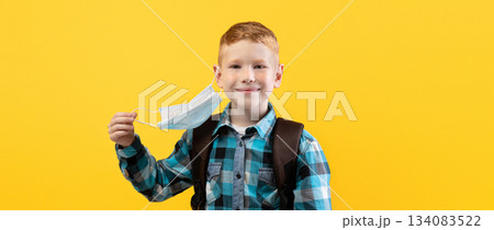 Smiling schoolboy taking off protective face mask over yellow studio background, copy space. Schoolboy holding face mask in hand, using protection against coronavirus while going to school Smiling schoolboy taking off protective face mask over yellow studio background, copy space. Schoolboy holding face mask in hand, using protection against coronavirus while going to school 134083522