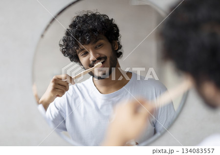Toothcare routine concept. Young indian guy cleaning teeth in the morning using toothbrush and toothpaste, standing near round mirror in bathroom 134083557