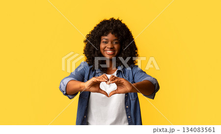 I love you. Happy african american young lady with curly hair showing heart shaped figure with both hands as a symbol of love, smiling at camera, yellow studio background, copy space I love you. Happy african american young lady with curly hair showing heart shaped figure with both hands as a symbol of love, smiling at camera, yellow studio background, copy space 134083561