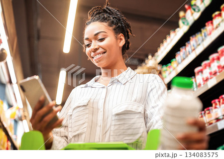 Happy Black Lady Using Cellphone With Grocery Shopping Checklist Application Buying Food Products In Supermarket Groceries Shop, Standing Wih Trolley Cart In Store. Selective Focus Happy Black Lady Using Cellphone With Grocery Shopping Checklist Application Buying Food Products In Supermarket Groceries Shop, Standing Wih Trolley Cart In Store. Selective Focus 134083575