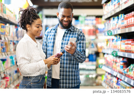 Black Family Doing Grocery Shopping In Supermarket. African American Couple Using Phone Scanning Food Product Standing In Store Aisles Indoors. Customers Buying Supplies In Local Shop 134083576