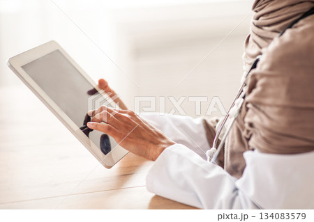 Cropped of female doctor in hijab using gadgets at workplace, holding digital tablet. Unrecognizable therapist muslim woman having online conversation with her patients, using pad, side view Cropped of female doctor in hijab using gadgets at workplace, holding digital tablet. Unrecognizable therapist muslim woman having online conversation with her patients, using pad, side view 134083579