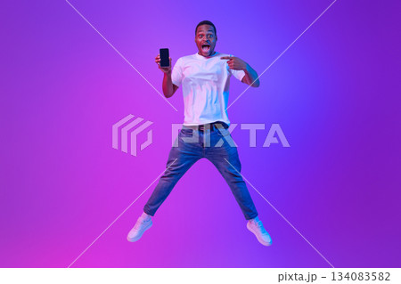 Portraif Of Amazed African Man Jumping Over Purple Neon Background And Pointing At Smartphone With Black Screen, Excited Guy Demonstrating Copy Space For Your Mobile Advertisement, Mockup Image Portraif Of Amazed African Man Jumping Over Purple Neon Background And Pointing At Smartphone With Black Screen, Excited Guy Demonstrating Copy Space For Your Mobile Advertisement, Mockup Image 134083582