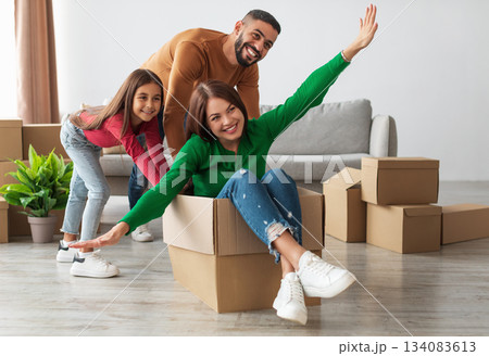 Celebrating Moving Day. Portrait of happy Arab family having fun together in new apartment, unpacking, smiling husband riding excited wife in cardboard box in living room with little daughter 134083613