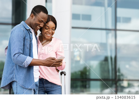 Cheerful black couple with smartphone booking hotel online after arrival to new country, romantic african american spouses standing near airport building, making accommodation reservation in internet 134083615