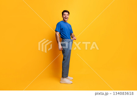 Successful Weight Loss. Happy Excited Slim Young Guy Pulling Old Large Loose Jeans Achieved Slimming Weightloss Goal Standing Posing Isolated On Yellow Studio Wall, Full Body Length, Free Copy Space 134083618