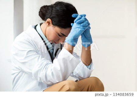 Stressed brunette young woman doctor having difficult day at clinic, wearing protective gloves, sitting next to wall in her office and resting, empty space. Upset female doctor feeling exhausted Stressed brunette young woman doctor having difficult day at clinic, wearing protective gloves, sitting next to wall in her office and resting, empty space. Upset female doctor feeling exhausted 134083659