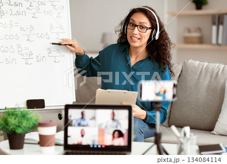 Videoconference And Training. Class of diverse students learning remotely online using web app. Female teacher in headset providing lesson looking at pc screen, recording video on cellphone on tripod 134084114