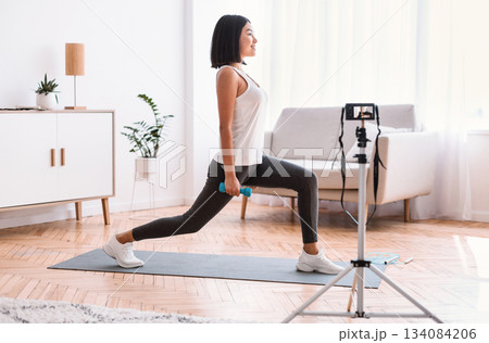 Online Workout Concept. Active asian lady filming home training tutorial on camera, doing lunges with dumbbells, exercising on yoga mat. Trainer recording video for internet channel in living room Online Workout Concept. Active asian lady filming home training tutorial on camera, doing lunges with dumbbells, exercising on yoga mat. Trainer recording video for internet channel in living room 134084206