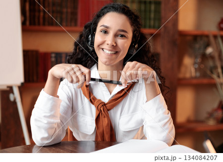 Front portrait of confident smiling curly young woman wearing headset sitting at desk at home office, library with bookshelves in the blurred background. Female student holding pen, posing at camera 134084207