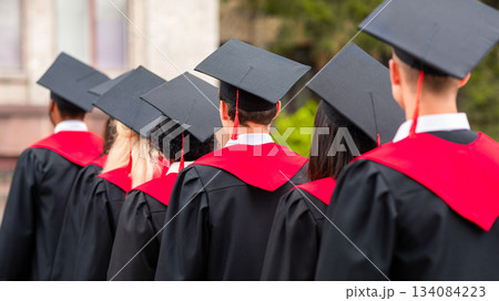 Back view of international group of students in graduation robes and caps walking by university campus, closeup. Unrecognizable multiracial students attending graduation ceremony 134084223