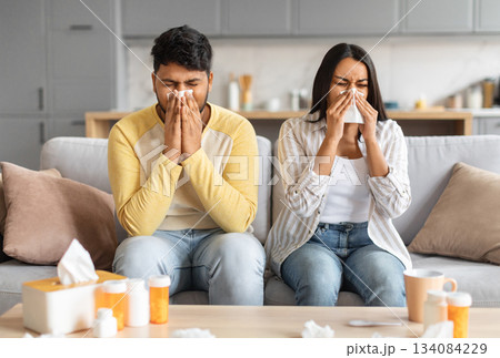 Sick indian couple sitting on sofa, blowing noses and suffering from colds, ill eastern spouses feeling unwell, relaxing on sofa in living room surrounded by medications, free space 134084229