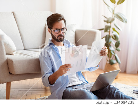 Busy Young Arab Entrepreneur Checking Papers And Using Laptop Computer At Home, Eastern Businessman Reading Financial Reports, Working Remotely, Enjoying Distance Job Opportunities, Free Space Busy Young Arab Entrepreneur Checking Papers And Using Laptop Computer At Home, Eastern Businessman Reading Financial Reports, Working Remotely, Enjoying Distance Job Opportunities, Free Space 134084239