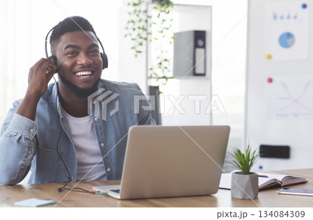 Continuing education program. Smiling african american employee in headphones watching webinar on laptop in modern office. Continuing education program. Smiling african american employee in headphones watching webinar on laptop in modern office. 134084309