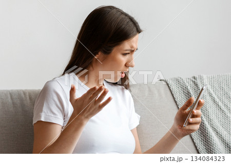 Stressed Young Pregnant Woman Looking At Smartphone Screen While Relaxing On Couch, Worried Expectant Lady Reading Unpleasant Message In Mobile Phone, Emotionally Reacting To Bad News, Free Space 134084323