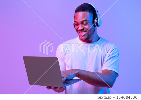 Online Gaming. Portraif Of Happy Black Man In Wireless Headphones Using Laptop Computer, Playing Games In Internet While Standing In Neon Lighting Over Purple Studio Background, Copy Space 134084330