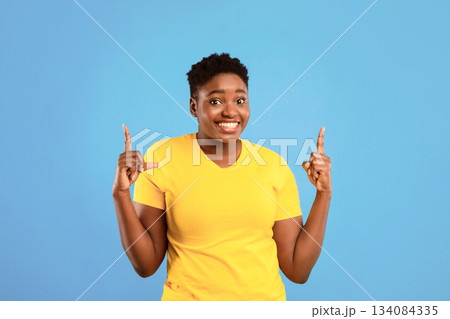 Look Upward. Cheerful African Plus-Size Lady Pointing Fingers Up Standing Posing On Blue Studio Background. Plump Black Female Showing Look There Advertising Something Upward 134084335