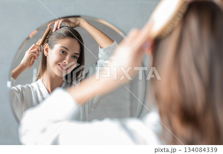 Portrait Of Young Attractive Lady Combing Her Hair With Brush And Looking At Mirror While Standing Near Mirror In Bathroom, Beautiful Female Enjoying Self-Care, Selective Focus On Reflection 134084339