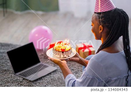 African woman in party hat celebrating birthday online at quarantine or self-isolation, using laptop with empty screen for video call with friends and family, holding cake, sitting on floor African woman in party hat celebrating birthday online at quarantine or self-isolation, using laptop with empty screen for video call with friends and family, holding cake, sitting on floor 134084343