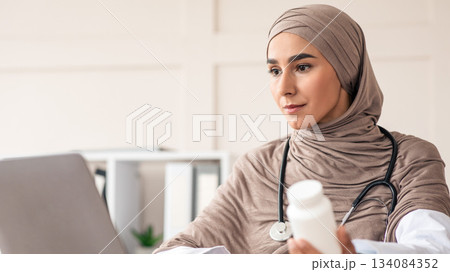 Closeup of female doctor young woman in hijab holding jar with medicine supplements or vitamins and using laptop, muslim lady physician working online from office in clinic, copy space Closeup of female doctor young woman in hijab holding jar with medicine supplements or vitamins and using laptop, muslim lady physician working online from office in clinic, copy space 134084352