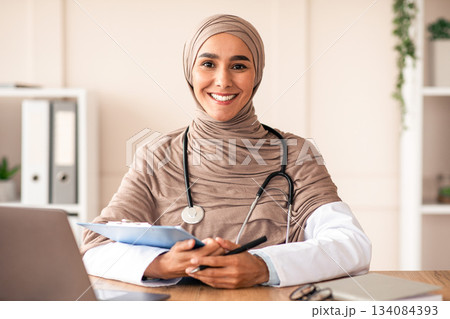Smiling young woman doctor in hijab taking anamnesis in hospital, sitting in front of laptop, holding medical chart and pen, copy space. Female therapist in head scarf smiling at camera while working 134084393