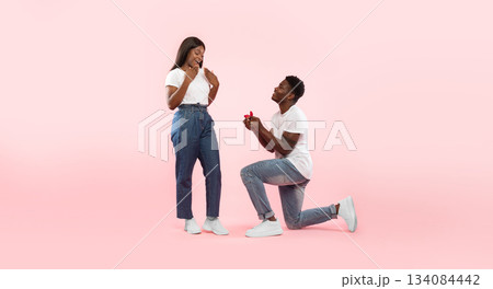 Marry Me. Happy black man holding giving open box with engagement ring to excited girlfriend asking her to be his wife during romantic date standing on one knee, pink studio wall, banner, copy space 134084442