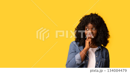 Interested pretty black woman in casual outfit looking at copy space and touching her chin, yellow studio background, panorama. Beautiful african american curly lady excited about advertising Interested pretty black woman in casual outfit looking at copy space and touching her chin, yellow studio background, panorama. Beautiful african american curly lady excited about advertising 134084479