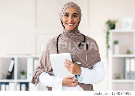 Modern technologies in healthcare concept. Happy female doctor in hijab posing with digital tablet in her office, copy space. Muslim woman therapist holding digital pad, clinic interior Modern technologies in healthcare concept. Happy female doctor in hijab posing with digital tablet in her office, copy space. Muslim woman therapist holding digital pad, clinic interior 134084493