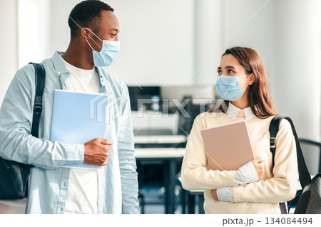 Education, Lifestyle And People Concept. Portrait of smiling diverse international students wearing protective medical masks and talking, standing in lecture hall at the university, wearing backpacks 134084494