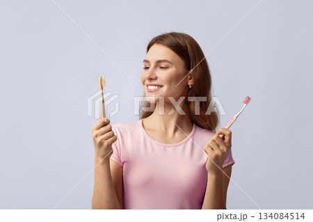 Smiling young woman holding eco bamboo and plastic toothbrush, choosing between different items for dental hygiene. Eco-friendly lady looking at wooden brush, standing on grey background, copy space Smiling young woman holding eco bamboo and plastic toothbrush, choosing between different items for dental hygiene. Eco-friendly lady looking at wooden brush, standing on grey background, copy space 134084514