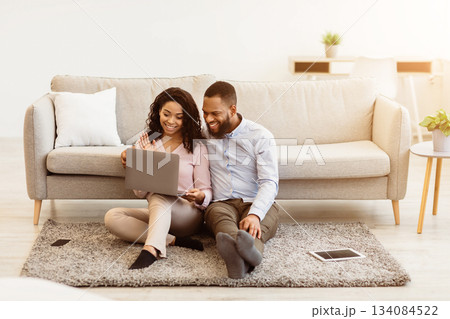 Virtual Communication. Happy black couple having online video conference with friends or family using computer, waving hand to web camera, sitting on the floor carpet in bedroom at home Virtual Communication. Happy black couple having online video conference with friends or family using computer, waving hand to web camera, sitting on the floor carpet in bedroom at home 134084522