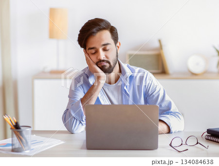 Overworked arab male entrepreneur sitting at desk in office and sleeping, eastern man napping at workplace, placed head on hand, feeling tired after working on laptop computer, copy space Overworked arab male entrepreneur sitting at desk in office and sleeping, eastern man napping at workplace, placed head on hand, feeling tired after working on laptop computer, copy space 134084638