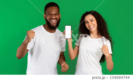 Diverse Couple Showing Cellphone Empty Screen Gesturing Yes Standing On Green Studio Background, Wearing White T-Shirts. New Great Application Concept. Panorama, Mockup 134084744