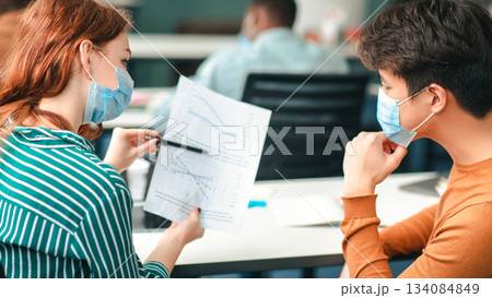 Healthcare, Education And People Concept. Group of diverse international students wearing protective medical masks and talking, discussing project, sitting at desk in lecture hall at the university Healthcare, Education And People Concept. Group of diverse international students wearing protective medical masks and talking, discussing project, sitting at desk in lecture hall at the university 134084849
