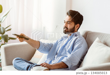 Arab Man Watching TV At Home. Young Eastern Guy With Remote Controller In Hand Resting In Living Room, Sitting On Couch And Switching Television Channels, Millennial Male Enjoying Domestic Rest Arab Man Watching TV At Home. Young Eastern Guy With Remote Controller In Hand Resting In Living Room, Sitting On Couch And Switching Television Channels, Millennial Male Enjoying Domestic Rest 134085013