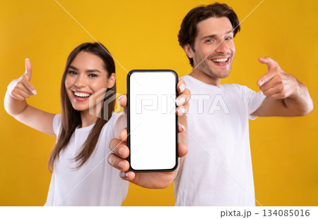 Use this New Application. Portrait of cheerful millennial couple pointing at smartphone with white screen, recommending mobile app, advertising your website, mock up for design. Selective focus 134085016