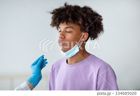 Coronavirus diagnostics. Doctor in rubber gloves making nasal coronavirus PCR test for African American teen, using sterile swab stick indoors. Black adolescent undergoing covid-19 testing procedure Coronavirus diagnostics. Doctor in rubber gloves making nasal coronavirus PCR test for African American teen, using sterile swab stick indoors. Black adolescent undergoing covid-19 testing procedure 134085020