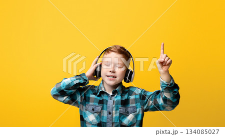 Cute redhead boy with closed eyes in wireless headphones listening to music and singing, aiming finger up and dancing, yellow studio background, copy space. Kids entertainment and lifestyle 134085027