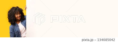 Amazed stylish african american lady in jeans shirt looking at camera, standing by white blank advertisement board for your text or announcement, yellow studio background, panorama 134085042