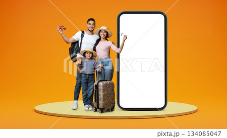 Smiling arabic family with small daughter enjoy vacation with suitcases, passports, car and plane on stage on orange studio background with huge phone with empty screen. App for trip, travel blog 134085047