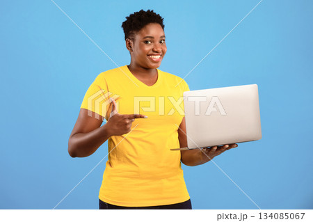 Oversized Black Woman Pointing Finger At Laptop Posing Holding Computer Advertising Website Standing In Studio Over Blue Background, Smiling To Camera. Online Service Ad Oversized Black Woman Pointing Finger At Laptop Posing Holding Computer Advertising Website Standing In Studio Over Blue Background, Smiling To Camera. Online Service Ad 134085067
