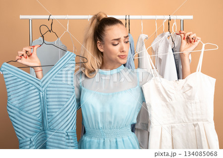 Making Choice. Portrait of young pensive woman holding two hangers with dresses and choosing trendy outfit, thinking about purchase and what to wear, standing in wardrobe, clothing rail in background Making Choice. Portrait of young pensive woman holding two hangers with dresses and choosing trendy outfit, thinking about purchase and what to wear, standing in wardrobe, clothing rail in background 134085068