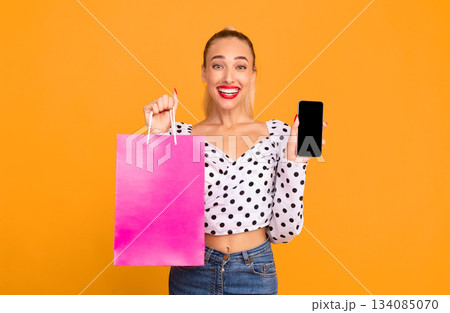 Application For Shopping Online. Portrait of excited woman showing cellphone with black empty screen for mock up to camera and holding pink paper bag, standing at pastel orange studio wall background 134085070