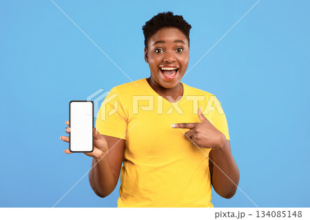 Excited Black Woman Pointing At Phone Blank Screen Showing Smartphone To Camera Recommending Application Over Blue Studio Background. Great Mobile App For Cellphone Concept. Mockup 134085148