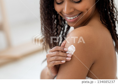 Body Care Concept. Smiling Beautiful Black Woman Applying Moisturizing Lotion On Shoulder, Happy African American Female Making Beauty Treatments While Standing Wrapped In Towel After Bath, Closeup 134085237
