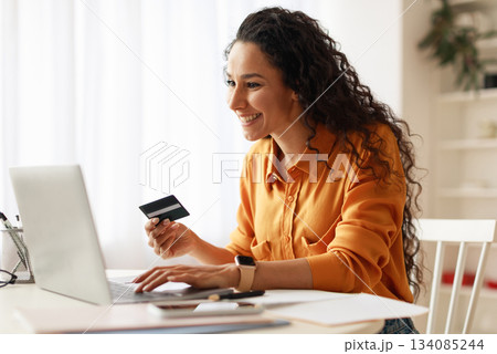 Happy Middle Eastern Woman Shopping Online Using Credit Card And Laptop Making Payment Sitting At Desk At Home. Internet Banking And Commerce, Shopaholism Concept. Side View 134085244